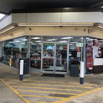 Shell - Entrance - The image shows a Shell gas station convenience store with a glass storefront, allowing visibility of shelves stocked with snacks, beverages, and other items. The entrance has signs indicating express lane and self-checkout options. Outside, there are trash bins, a trash receptacle, and yellow-striped parking or loading zones on the concrete pavement. Promotional posters, including Coca-Cola advertisements, are displayed on the exterior walls. The store is situated under a canopy, with trees visible in the background. - Gas Stations Near Me - Hoover, Alabama
