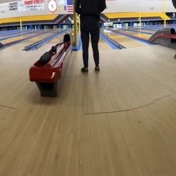 Bowling Alleys - A spacious indoor bowling alley features multiple lanes with wooden flooring, red ball return systems, and several people standing or walking near the lanes. The background includes colorful wall decorations and a digital scoreboard. The environment appears well-lit and designed for recreational bowling activities.