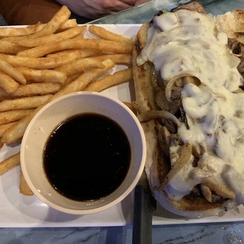 Kelly's Bleachers - Philly cheese steak sandwich. - A Philly cheesesteak sandwich topped with melted cheese and sautéed onions, served with a side of French fries and a small bowl of dark dipping sauce. - Sports Bars Near Me - Milwaukee, Wisconsin