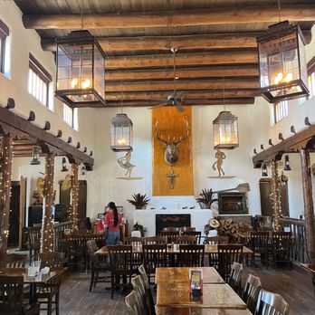 Frankie's at the Cassanova - the dining room - The image shows the interior of a rustic-style restaurant featuring wooden beams on the ceiling and wooden flooring. Hanging lanterns provide lighting, and there are small windows along the upper walls. A mounted deer head with antlers is centered on the wall behind a fireplace, flanked by decorative figures and potted plants. Several tables and chairs are arranged throughout the space, with a person standing near one of the tables. The decor emphasizes a lodge or cabin aesthetic. - New American Near Me - Pecos, New Mexico
