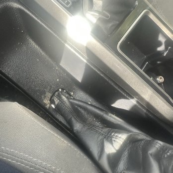 Riverhead Auto Wash - The photo captures the interior of a vehicle, highlighting the gear shift lever and surrounding console area. A person's hand, wearing a black leather glove, is resting near the gear shift. The gear shift is in the neutral position, and the console features a small storage compartment. The image is taken from a close angle, with sunlight reflecting off the gear shift area, and the surrounding surfaces are black and textured. - Auto Detailing Near Me - Riverhead, New York