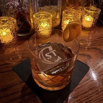 Constitution Bar - A cocktail with a lemon twist garnish is placed on a black coaster on a wooden bar surface. The background features glass jars with candles inside, creating a warm, dimly lit ambiance. The setting suggests a cozy bar environment, possibly a speakeasy-style venue with decorative candle holders and a focus on craft cocktails. - Speakeasies Near Me - Lexington, Kentucky
