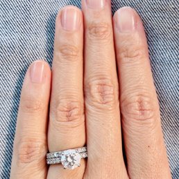 A close-up of a hand wearing an engagement ring with a large central diamond and smaller diamonds on the band, resting on a fabric surface.