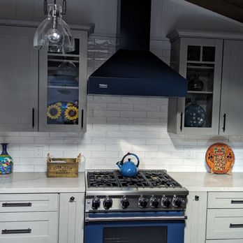 Dan Construction - The kitchen has a black range hood mounted above a blue stove with a kettle on top. The backsplash is made of white subway tiles, and the countertop holds decorative items including a sunflower vase, a wooden box, and a colorful plate. The cabinetry is light gray with black handles, and the ceiling is wood-paneled. - General Contractors Near Me