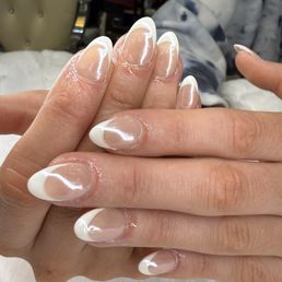 Hands with clear, glossy nails featuring French tips, resting on a surface, likely in a nail salon setting.