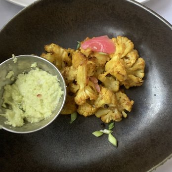 The Fat Radish - Curried cauliflower - Roasted cauliflower florets served on a black plate, accompanied by a small metal cup of mashed potatoes and garnished with sliced green onions. - Breakfast & Brunch Near Me - Cornucopia, Wisconsin