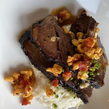 The Kitchen Table - A dish featuring braised beef short ribs with a dark, caramelized exterior and tender interior is presented on a white plate. The ribs are accompanied by a portion of mashed potatoes and topped with a corn relish that includes diced tomatoes and chopped chives. The presentation emphasizes the hearty, savory nature of the meal, suitable for a comfort food setting. - American Near Me - Clermont, Kentucky