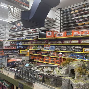Trainland - The image shows a model train layout on a table in a retail store, with tracks, bridges, and miniature scenery. Shelves on the walls are stocked with boxed model trains and accessories in various scales and brands. The store has bright lighting and organized displays, indicating a large hobby shop specializing in model trains. - Toy Stores Near Me - Lynbrook, New York