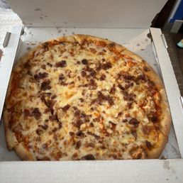 bacon chicken buffalo ranch - A pizza with a golden crust topped with melted cheese, bacon bits, chicken pieces, and drizzled with buffalo sauce and ranch dressing, inside an open white pizza box. The box is on a surface with part of a wall and some furniture visible in the background.