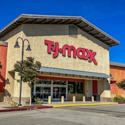 The image shows the exterior of a TJ Maxx retail store located in a shopping center. The building features a beige facade with red accents and large, prominent red signage displaying the store's name. The entrance has glass doors and windows, with a small awning above. The parking lot in front is mostly empty, with a few small trees and street lamps visible. The sky is clear and blue, indicating a sunny day.