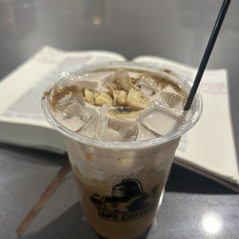 Ape Coffee - Banana latte - A banana latte with ice cubes and toppings in a clear plastic cup, placed on a gray table with an open book in the background. - Coffee & Tea Near Me - Orange, California