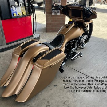 Straightline Custom's - A matte gold custom motorcycle with a sleek, elongated design is parked on a concrete surface at a gas station. The motorcycle features large, streamlined saddlebags and a black seat, with a digital display and handlebars visible. The background includes a brick wall, a fuel pump, and vehicles, indicating an outdoor setting. There is a humorous caption about the motorcycle's appearance and a critique of a person named John. - Motorcycle Repair Near Me - Mesa, Arizona