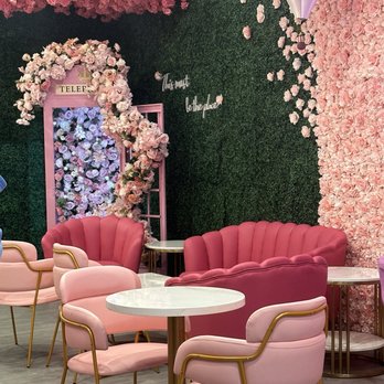 Bober Tea - Beautiful! - The image shows a seating arrangement with pink upholstered chairs and sofas, set against a backdrop of floral wall decor and greenery. There is a hot air balloon decoration hanging from the ceiling, and the walls are decorated with pink and white flowers. A floral arch and a pink telephone booth-like structure with flowers are also visible, creating a decorative and themed ambiance. - Donuts Near Me - Carmel, Indiana