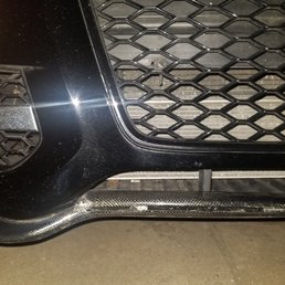 My cracked Carbon Fiber lip spoiler TNS Auto Group damaged. - The image depicts the front section of a black car, focusing on the grille and bumper area. The grille has a honeycomb pattern, and the bumper shows visible scratches and scuffs. A carbon fiber trim is present at the lower part of the bumper, and the surface appears to have some dirt or dust. The lighting highlights the reflective surface of the car's paint and the textured pattern of the grille.