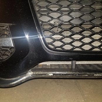 TNS Auto Collision - My cracked Carbon Fiber lip spoiler TNS Auto Group damaged. - The image depicts the front section of a black car, focusing on the grille and bumper area. The grille has a honeycomb pattern, and the bumper shows visible scratches and scuffs. A carbon fiber trim is present at the lower part of the bumper, and the surface appears to have some dirt or dust. The lighting highlights the reflective surface of the car's paint and the textured pattern of the grille. - Body Shops Near Me - Brooklyn, New York