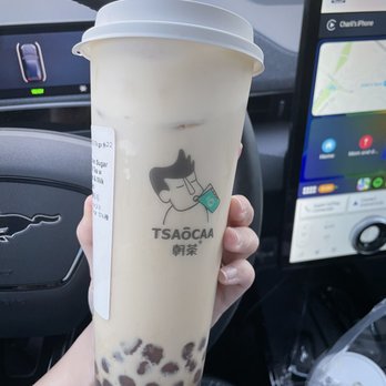 Tsaocaa - Austin - $6.75 Brown Sugar Bubble Milk Tea with Oreo and Milk Foam / - A hand holds a large bubble tea cup with tapioca pearls at the bottom inside a vehicle, with a car dashboard and navigation screen in the background. - Bubble Tea Near Me - Austin, Texas