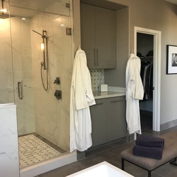 Tru Builders - AFTER - Shower & cabinet - The image shows a contemporary bathroom with a glass shower enclosure, marble tile walls, and a tiled shower floor. Adjacent to the shower are gray cabinets with silver handles, and a small countertop with a tissue box. Two white bathrobes hang on hooks on the cabinet doors. A bench with folded dark towels is positioned on the dark tiled floor. An open closet with hanging clothes and a framed black-and-white photograph on the wall are also visible. - General Contractors Near Me - Surprise, Arizona