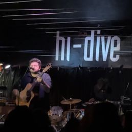A live music performance at Hi-Dive in Denver features a guitarist singing into a microphone, with drums and a DJ setup visible in the background.