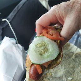 Jumbo Vienna Chicago style hot dog - A hand is holding a hot dog with cucumber slices, tomato, and other toppings, wrapped in paper, inside a vehicle. The background shows the car's interior, including a seat and a window with greenery outside.