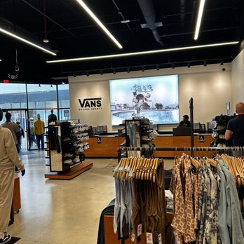 Vans - The image shows the interior of a Vans retail store located in Orange County. Clothing racks with various apparel are positioned in the foreground, and the store features a modern design with black ceiling panels and linear lighting. A large digital screen displaying an image is mounted on the back wall, alongside the Vans logo and the text "Orange County." Several customers are browsing, and the entrance with glass doors is visible on the left side of the image. - Shoe Stores Near Me - Orange, California