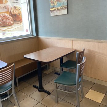 McDonald's - Dirty Table - A corner of a restaurant features a wooden table with a black metal base, two chairs with green seats and wooden backs, and a built-in bench with a brown cushion. The wall behind has a textured blue-green finish with a small painting of trees. A large window displays an advertisement for donuts, showing images of glazed and sprinkled donuts. The floor is tiled with beige and brown squares, and the space appears clean and quiet. - Burgers Near Me - Bourbonnais, Illinois