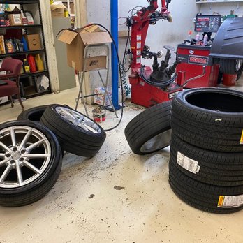 Q Auto Service - Four tires, including alloy wheels and plain tires, are stacked and laid on the floor of an auto repair shop with tools and shelves in the background. - Auto Repair Near Me - Springfield, Virginia