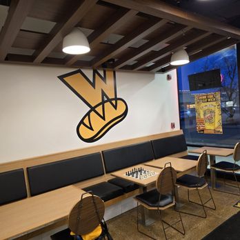 Which Wich - A sandwich shop interior features a wall with a logo, wooden and black seating, and a window with promotional posters. - Sandwiches Near Me - Arlington Heights, Illinois