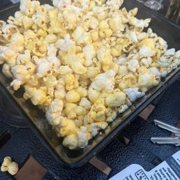 Free popcorn - A rectangular black tray contains a large serving of popcorn, with some pieces spilling over the edge. The tray is on a black metal table, and a glass and a drink container are visible behind it. There is also a paper label with handwritten text on the table.