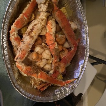 Costco - king crab legs feast - The image shows a foil tray filled with cooked crab legs, shrimp, and assorted vegetables, including corn on the cob. The seafood appears seasoned and ready to serve, suggesting a seafood feast or meal. The tray is placed on a surface with a green cloth nearby, and part of a chair and other household items are visible in the background. - Wholesale Stores Near Me - Brandywine, Maryland