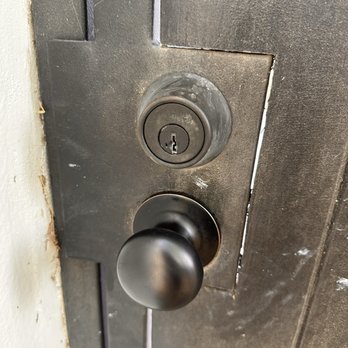 Desert Locksmith - A close-up view of a dark wooden door featuring a black doorknob and a deadbolt lock above it. The door frame is visible on the left side, with some minor wear and tear around the edges. The lock and knob are centered in the image, with the lock showing signs of use and the door surface appearing slightly textured. - Keys & Locksmiths Near Me