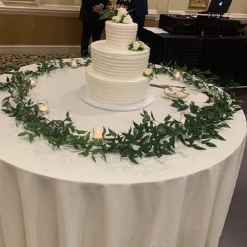 AGS Event Creations - A three-tiered white wedding cake with textured icing is positioned on a round table draped with a white tablecloth. The table is decorated with a circular arrangement of green leaves and small lit candles. Two men in formal suits are visible in the background, along with a DJ setup and a laptop. The setting appears to be an indoor event space with ornate wall decorations. - Wedding Planning Near Me - Mundelein, Illinois