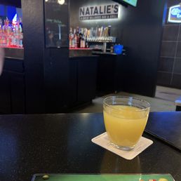 Bar - A beverage in a clear glass is placed on a coaster on a black bar table at Natalie's billiards bar and grill, with a bar area and screens visible in the background.