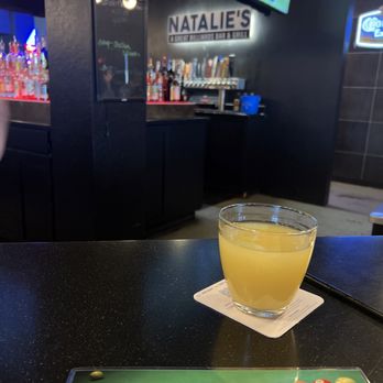 Natalie's - Bar - A beverage in a clear glass is placed on a coaster on a black bar table at Natalie's billiards bar and grill, with a bar area and screens visible in the background. - Sports Bars Near Me - Gilbert, Arizona