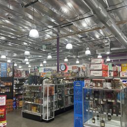 The image shows the interior of a retail store, likely a grocery or specialty shop, with multiple shelves and display cases filled with different products. The ceiling features exposed ductwork and numerous ceiling lights providing bright illumination. There are signs and packaging visible on the shelves, and the store appears organized with clear pathways for customers. The overall setting suggests a commercial retail environment focused on food or beverage items.