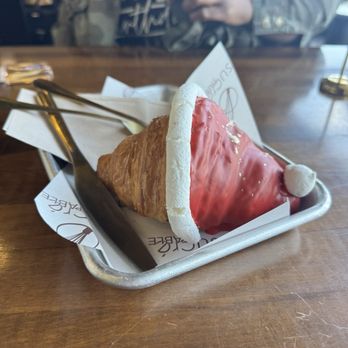 Sucre Table - A pastry shaped like a croissant is decorated with red icing covering part of it, topped with a small white fondant ball and white fondant trim resembling a Santa hat. It is served on a metal tray lined with branded paper, on a wooden table in a casual dining setting. A person in a jacket is visible in the background, holding a drink. - Bakeries Near Me - Tampa, Florida