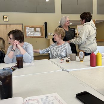 Open Door Cafe - High school reunion - Four women are seated at a rectangular table in a casual dining environment. One woman in a striped shirt reaches out to touch the arm of another woman with glasses and a gray sweater. Two other women are seated nearby, one with glasses and a gray shirt, and another partially visible. The table has condiments, a menu, and drinks. The background features a beige wall with framed items and acoustic panels. - Pizza Near Me - Lucas, Ohio