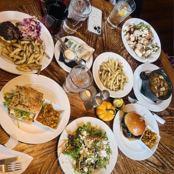 Roots Cafe - Our lunch - A round wooden table holds multiple plates of food, drinks, and a small pumpkin decoration, with a smartphone placed on the table. - New American Near Me - Naples, New York