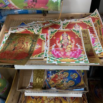 Double Fresh - The image shows a store shelf with various Hindu religious posters and images. Prominent among them are colorful depictions of Hindu deities, including Lakshmi sitting on a lotus flower, and other divine figures. The posters are arranged in a slightly overlapping manner, with some placed in a wire basket and others laid flat. The background includes a wooden surface and some additional items, such as balloons and packaging, indicating a retail environment for religious or cultural items. - International Grocery Near Me - Pittsburgh, Pennsylvania