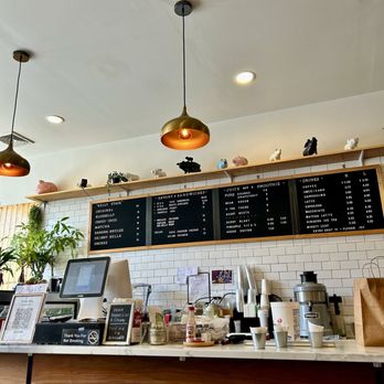 Belly Cake - The image shows the interior of a café with a white ceiling and a wooden counter. Two pendant lights hang from the ceiling. Behind the counter, black menu boards list various food and beverage options, including sandwiches, smoothies, and drinks. A shelf above the menu holds decorative pig figurines and small plants. The counter has a digital register, cups, condiments, and a large coffee machine. The overall setting is bright and modern, with a clean and organized appearance. - Pancakes Near Me - Bayside, New York