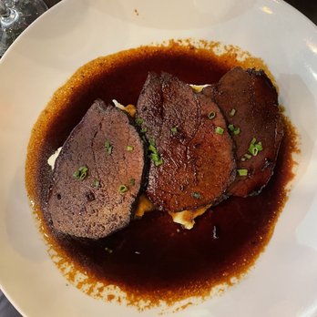 Bienmesabe - Three slices of cooked beef with a seared exterior are arranged on a white plate, topped with chopped green herbs. The beef is surrounded by a dark sauce that pools around the slices, indicating a flavorful preparation. The plate is set on a dark table, with a glass of water and a wine glass visible in the background, suggesting a dining setting. - Venezuelan Near Me - Chicago, Illinois