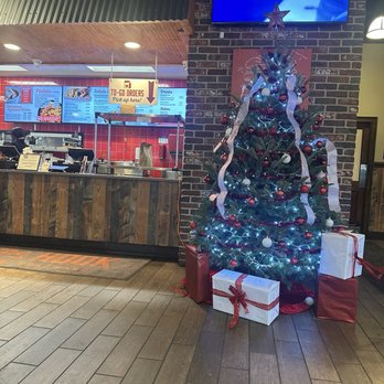 Off The Hook - A Christmas tree decorated with lights, ornaments, and ribbons is positioned against a brick wall in a restaurant, with wrapped gifts at its base. - Cajun/Creole Near Me - Hammond, Louisiana