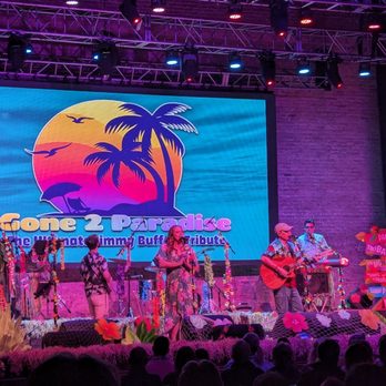 Arcada Theatre - a band performing on stage - A musical group performs on stage in front of an audience, with a large digital screen behind them showing a sunset scene with palm trees and birds, along with the text 'Gone 2 Paradise.' The stage is decorated with tropical-themed props, including flowers and signs pointing to various destinations. The performers include singers and guitarists, with stage lighting casting a colorful glow over the scene. Audience members are visible in the foreground, watching the performance. - Performing Arts Near Me - St. Charles, Illinois