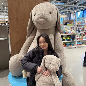 Indigo - A woman with long dark hair is sitting on a circular platform in a toy store, holding a small plush bunny. Behind her is a large plush bunny with floppy ears, sitting on the same platform. The store features shelves filled with toys, including cars and colorful items, with bright lighting and a modern interior design. The woman is smiling and dressed in a black jacket. - Bookstores Near Me - Toronto, Ontario
