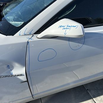 Body Master - The image depicts a white car with visible dents and scratches on the front fender and door. Handwritten notes and circles are marked on the car's surface, likely indicating areas needing repair or inspection. The side mirror has notes referencing the mirror and the door, with additional markings on the windshield and body panels. The car is parked on a concrete surface, and the photo appears to be taken outdoors in daylight. - Body Shops Near Me - Albuquerque, New Mexico