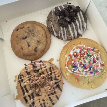 Dirty Dough - Dirty Four- Stuffed chocolate cake, sugar cookie, cookies and cream, The Reversal - A white bakery box holds four cookies, each with distinct toppings and decorations. The top left is a chocolate chip cookie with visible chocolate chips. The top right is a cookies and cream cookie topped with crushed Oreos and white icing drizzle. The bottom left is a peanut butter cookie with chocolate drizzle and crumbled topping. The bottom right is a sugar cookie decorated with colorful rainbow sprinkles and white icing drizzle. The cookies are arranged in a 2x2 grid inside the box. - Desserts Near Me - Kyle, Texas