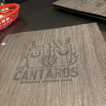 Los Cantaros - Menus - A menu with the Cantaros logo and text is placed on a wooden table at a Mexican restaurant, with another menu and a red basket nearby. - Mexican Near Me - Hampton, New Hampshire