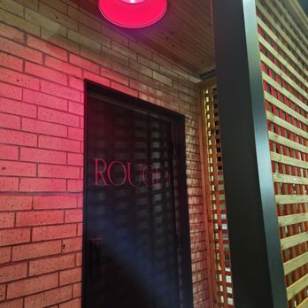 Rouge 41 - Red light above the door in the back - The image shows a black door with the word 'ROUGE' written on it, set into a brick wall. Above the door, a pink ceiling light provides illumination. To the right, there is a wooden lattice structure and a metal support beam, indicating an interior space with modern design elements. The ceiling is made of wood, adding warmth to the setting. - Speakeasies Near Me - Chaska, Minnesota