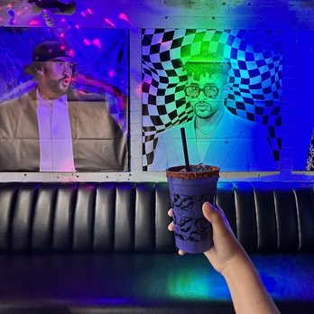 Cobra Arcade Bar - Benito themed drink! - A person’s hand is visible holding a drink with a salted rim and a black straw, positioned in front of a black leather bench. The background features two large, colorful posters with psychedelic and artistic designs, illuminated by multicolored lights, indicating an arcade bar setting. The environment includes vibrant lighting and decorative artwork, creating a lively atmosphere. - Bars Near Me - Phoenix, Arizona