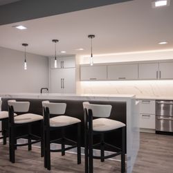 Finished Basements Plus - Featuring waterfall countertop, custom kitchen lighting, bar seating, white cabinetry, and more in this luxurious basement kitchen space