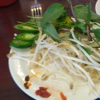 Pho Bar - They bring out a this plate with bean sprouts, fresh cilantro, basil and sliced jalapenos. - A white plate holds bean sprouts, fresh herbs, sliced jalapenos, and small amounts of sauces, set on a dark wooden table. - Vietnamese Near Me - Rio Rancho, New Mexico
